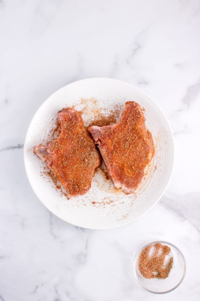 Raw pork chops being seasoned on a white dinner plate.
