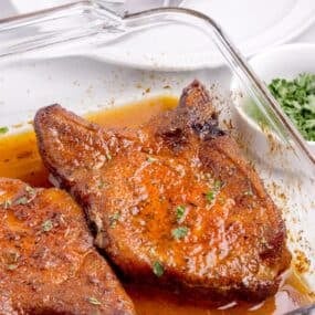 A glass baking dish with two baked pork chops with a sprinkle of parsley.