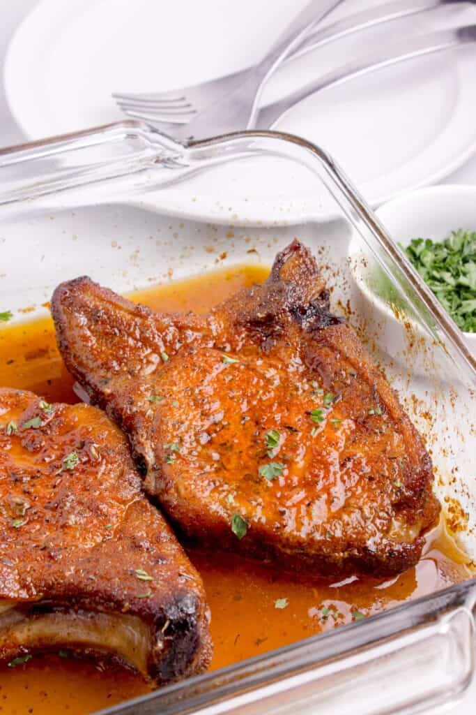 A glass baking dish with two baked pork chops with a sprinkle of parsley.