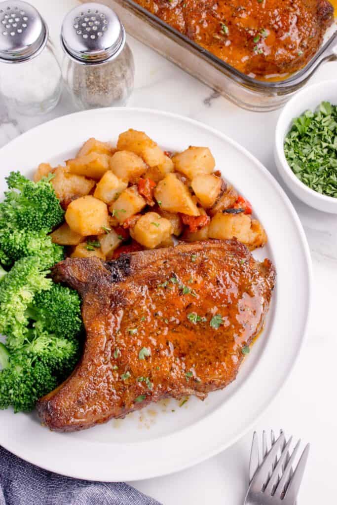 Pork chop on a white dinner plate with potatoes and broccoli. A small bowl of chopped parsley and glass baking pan is next to the plate with more baked pork chops.