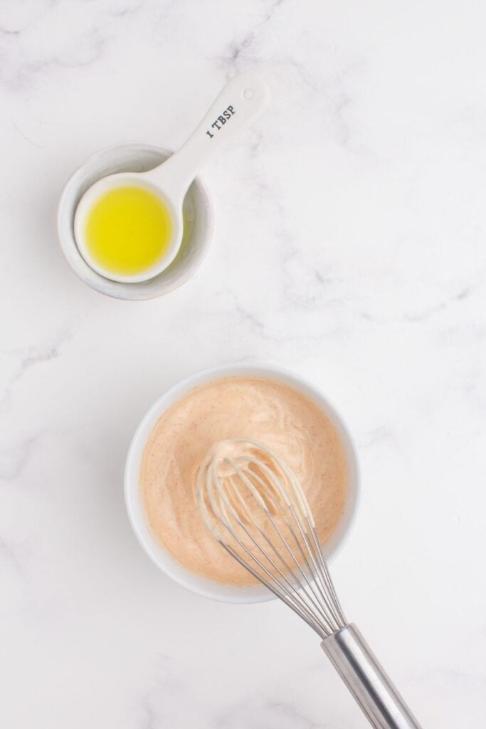 A small white bowl with ingredients for smash burger sauce being whisked together. A tablespoon of oil is next to the bowl.