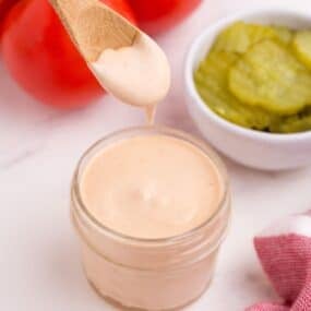 Hamburger sauce in a glass jar, surrounded by tomatoes on the vine and a bowl of pickles. A wooden spoon is lifting some sauce from the glass jar.