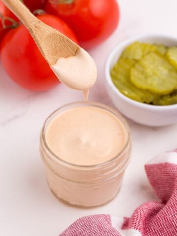 Hamburger sauce in a glass jar, surrounded by tomatoes on the vine and a bowl of pickles. A wooden spoon is lifting some sauce from the glass jar.