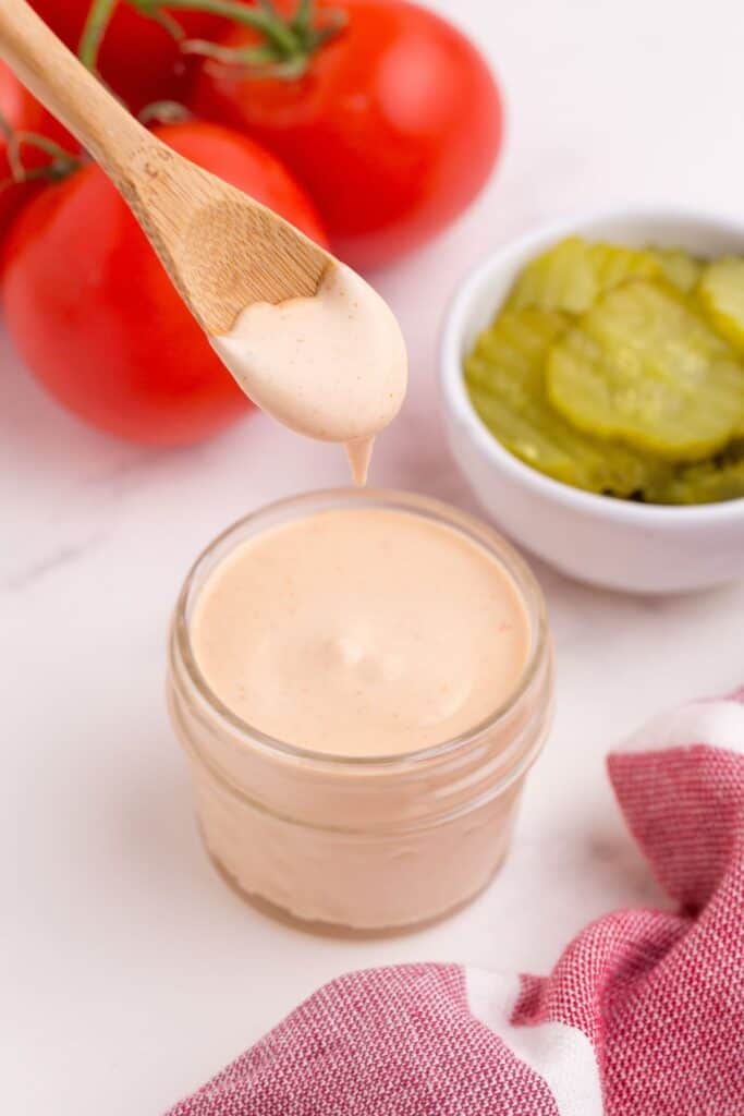 Hamburger sauce in a glass jar, surrounded by tomatoes on the vine and a bowl of pickles. A wooden spoon is lifting some sauce from the glass jar.