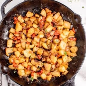 Cast iron pan with potatoes. A white dinner plate with forks and small bowl of parsley are above the pan.
