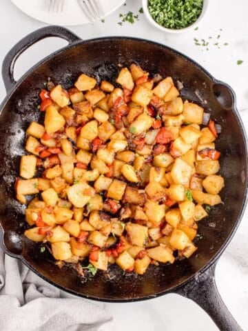 Cast iron pan with potatoes. A white dinner plate with forks and small bowl of parsley are above the pan.