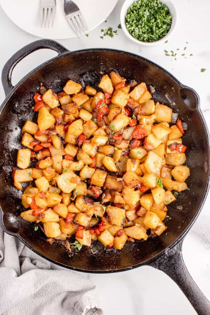 Cast iron pan with potatoes. A white dinner plate with forks and small bowl of parsley are above the pan.