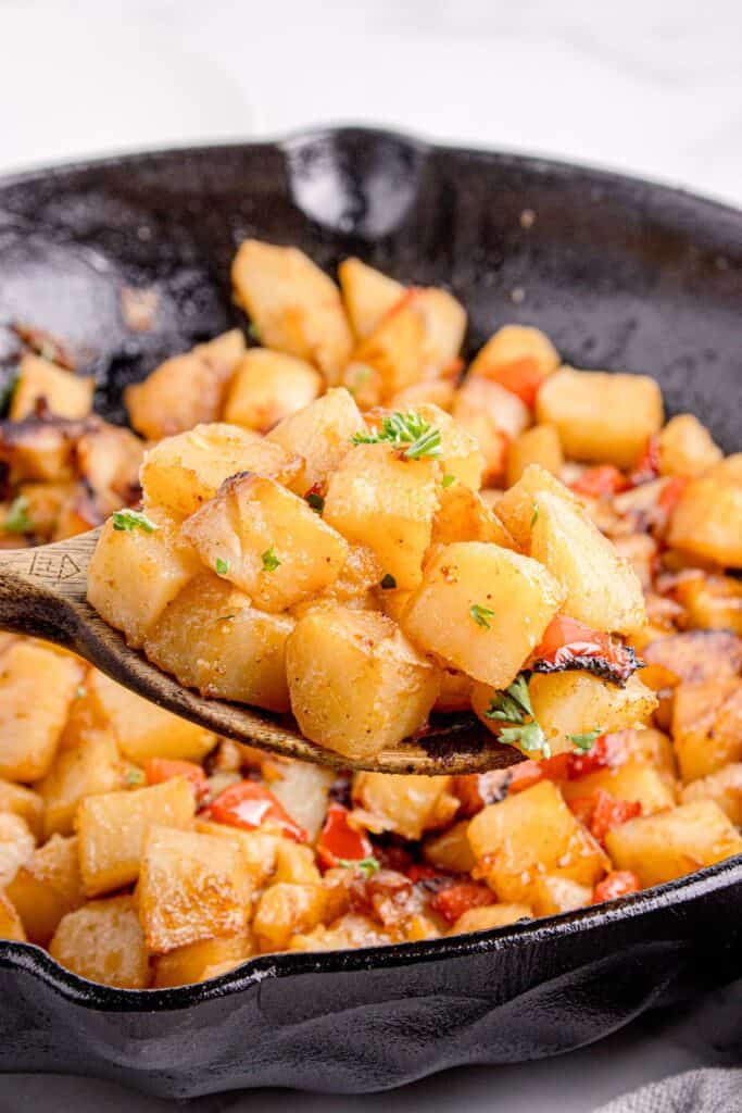 Chopped smothered potatoes in a cast iron pan. A wooden spoon is lifting a serving out of the pan.