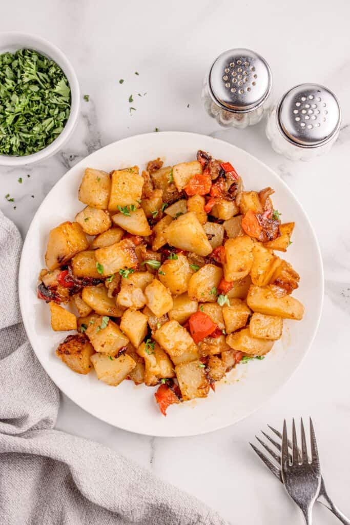 Completed smothered potatoes recipe on a white dinner plate surrounded by a small bowl of chopped parsley, forks, salt and pepper shakers.