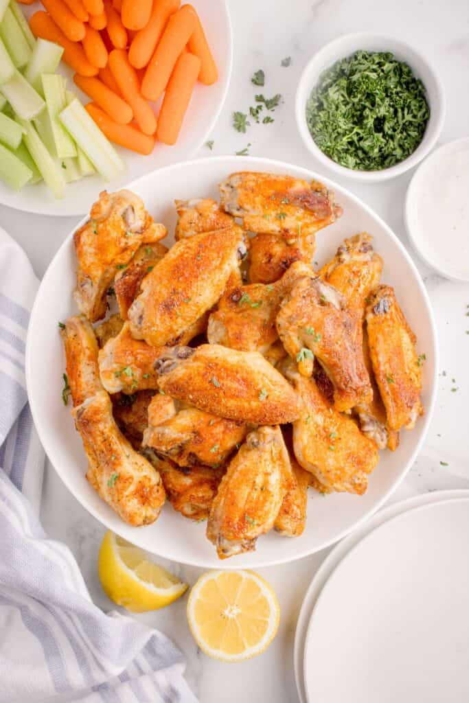 Pile of chicken wings on a white platter surrounded by a small bowl of dip, bowl of parsley, plate of carrots and celery, a cut lemon, and a stack of empty dinner plates.