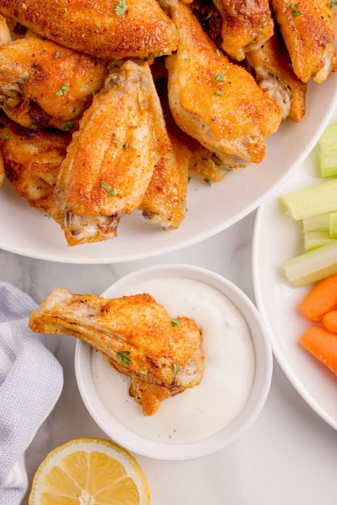 Platter of chicken wings next to a small bowl of dip and a plate of carrots and celery. A single chicken wing is placed in the dip.