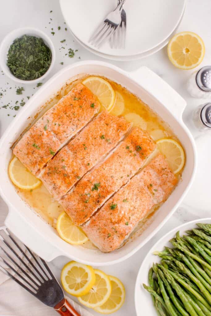 Four salmon filets in a white baking dish surrounded by a small bowl of parsley, stacked dinner plates, sliced lemons, a plate of asparagus, and a set of salt and pepper shakers.