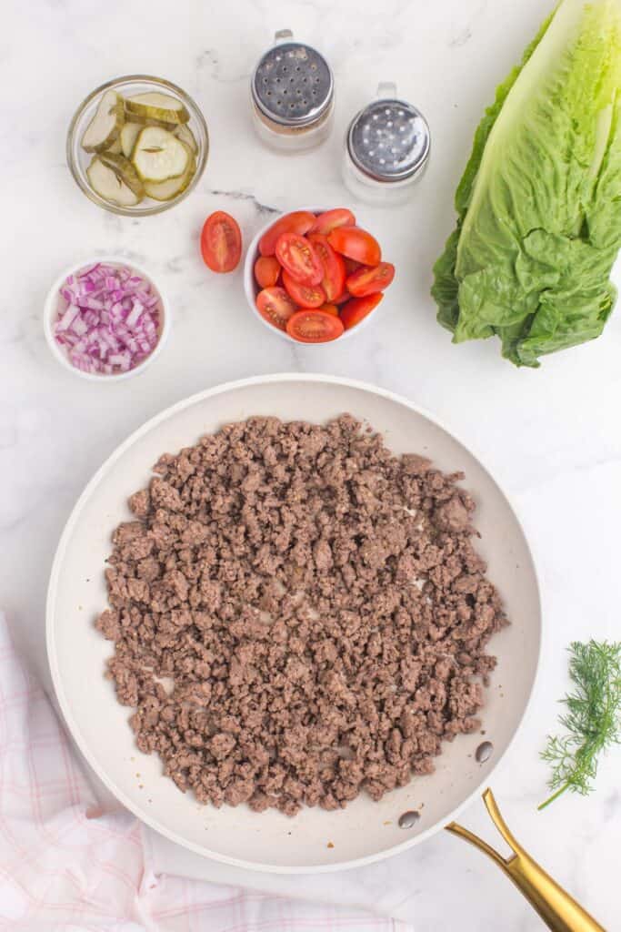 Cooked ground beef in a skillet next to prepped ingredients for burger bowls.