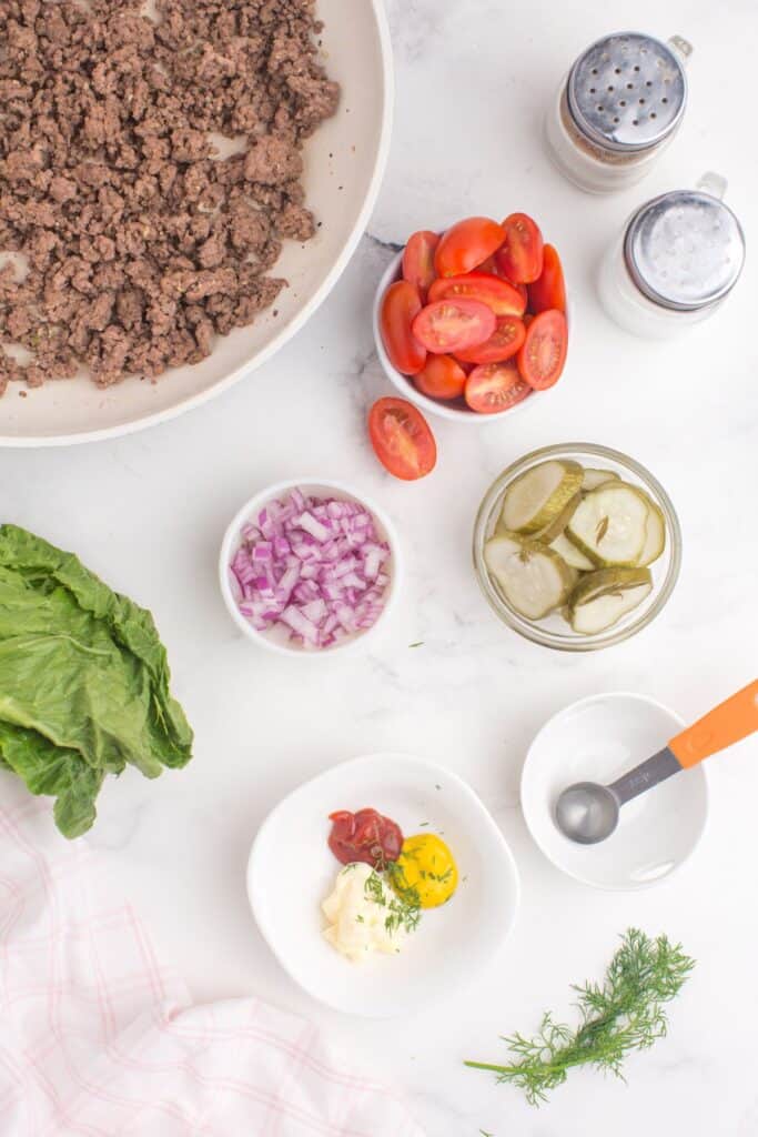 A skillet of cooked ground beef surrounded by small bowls of chopped purple onion, pickles, halved tomatoes, burger sauce ingredients, lettuce, and salt and pepper shakers.