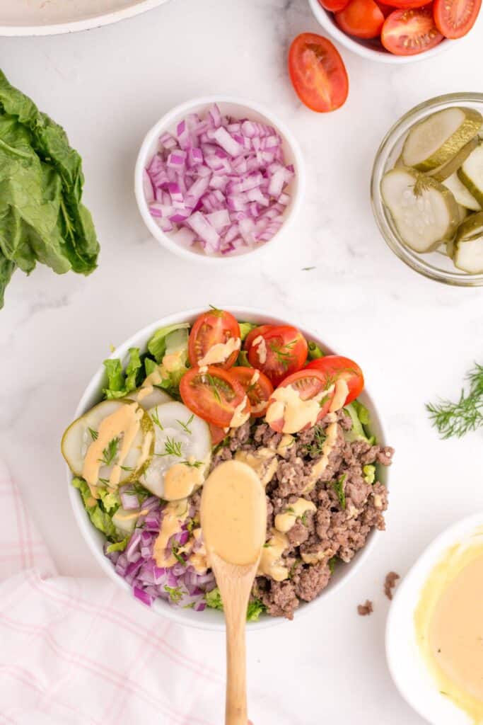 A wooden spoon drizzling burger sauce over the top of a burger bowl. Small bowls of burger sauce, pickles, chopped purple onion, and halved tomatoes are scattered around.