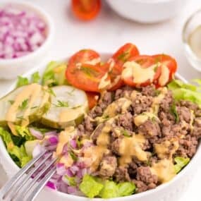 Burger bowl with burger sauce drizzled on top. Bowl is surrounded by small bowls of halved tomatoes and chopped purple onion.
