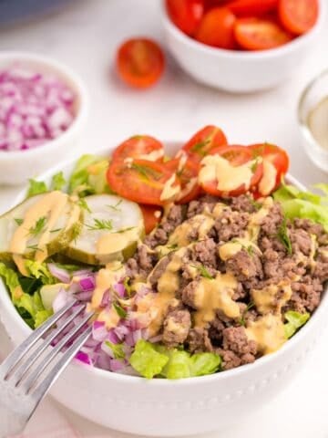 Burger bowl with burger sauce drizzled on top. Bowl is surrounded by small bowls of halved tomatoes and chopped purple onion.