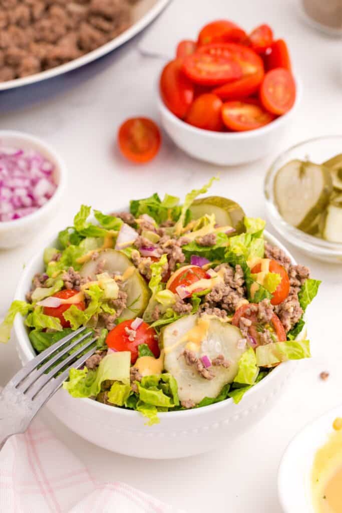 Completed burger bowl surrounded by small bowls of pickles, halved tomatoes, purple onion, and a skillet of ground beef.