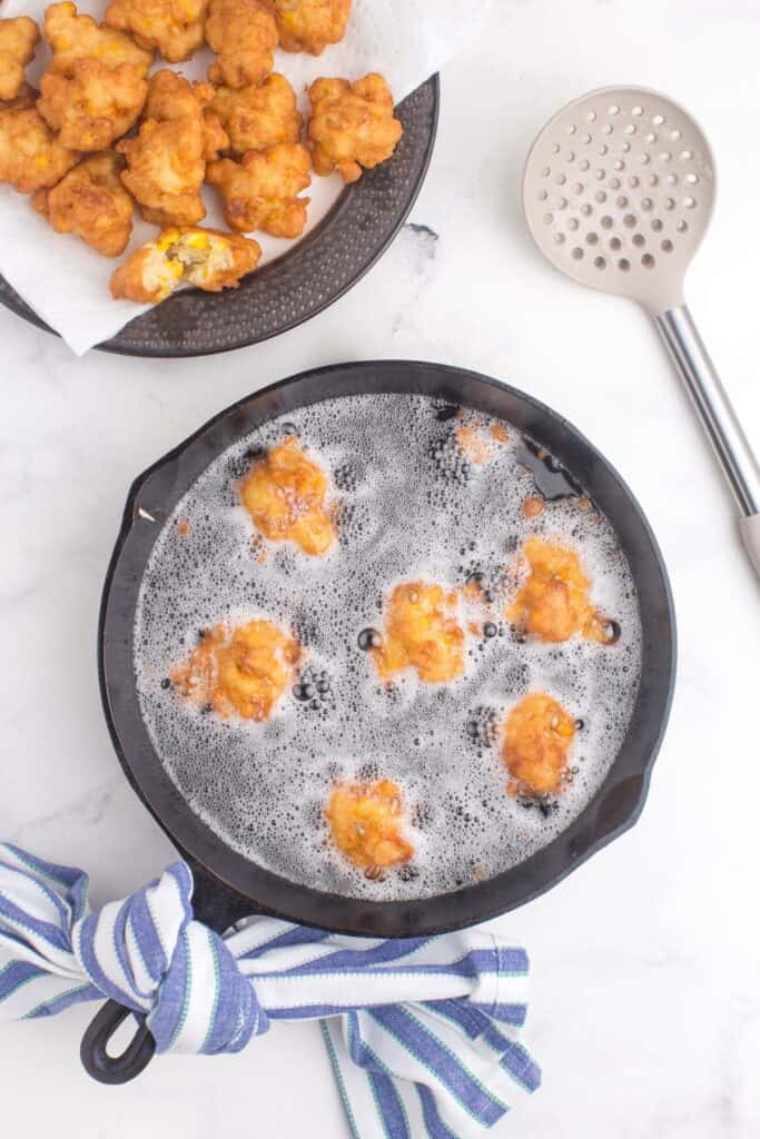 Cast iron pan with oil in it, frying corn nuggets. A strainer spoon is next to the pan. A serving platter is positioned next to the pan, with corn nuggets cooling on it.