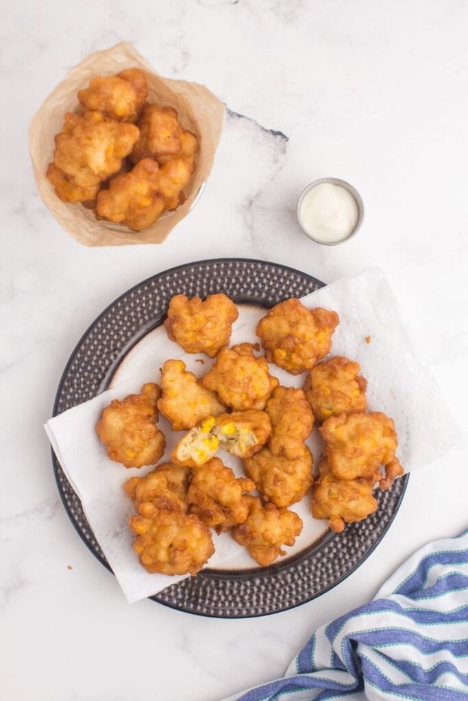 Gray serving platter lined with a white paper towel. Corn nuggets are piled up on the paper towel. Next to the platter is a fry stand filled with nuggets.