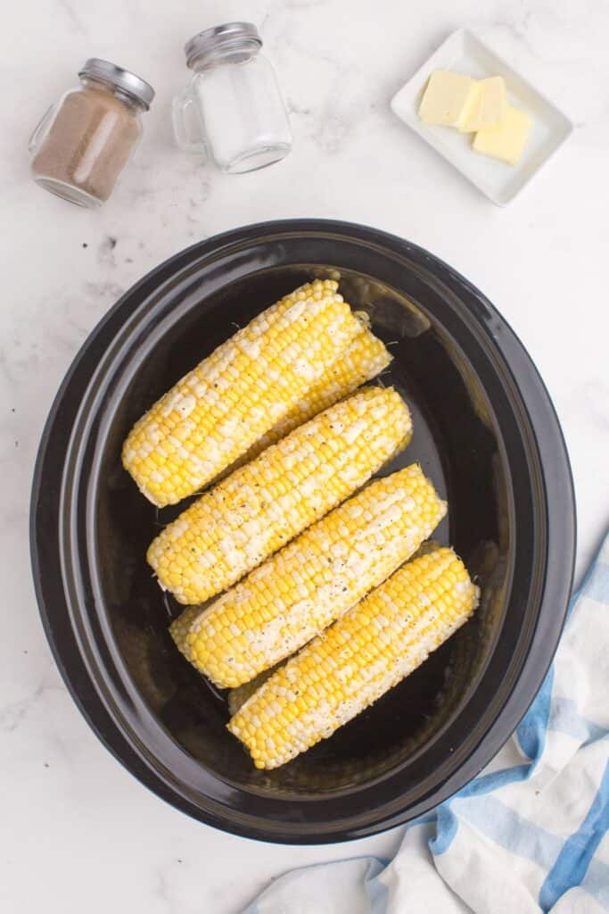 Uncooked ears of corn prepped and stacked in a crock pot insert. A small dish of butter and a set of salt and pepper shakers are above the insert.
