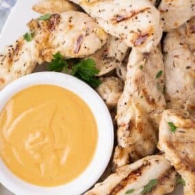 Chicken tenders garnished with parsley on a large dinner plate with a dipping sauce in a small bowl.