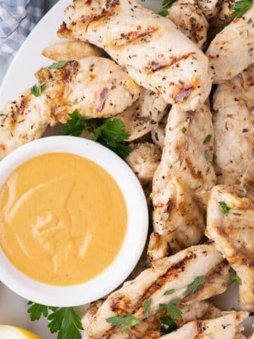 Chicken tenders garnished with parsley on a large dinner plate with a dipping sauce in a small bowl.