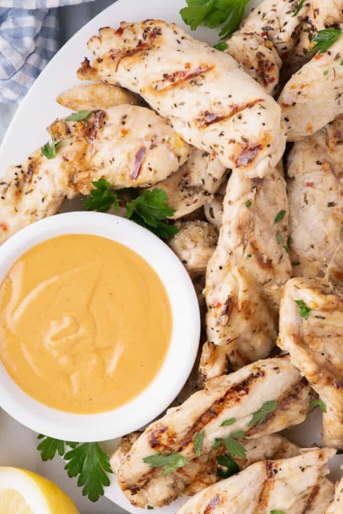 Chicken tenders garnished with parsley on a large dinner plate with a dipping sauce in a small bowl.