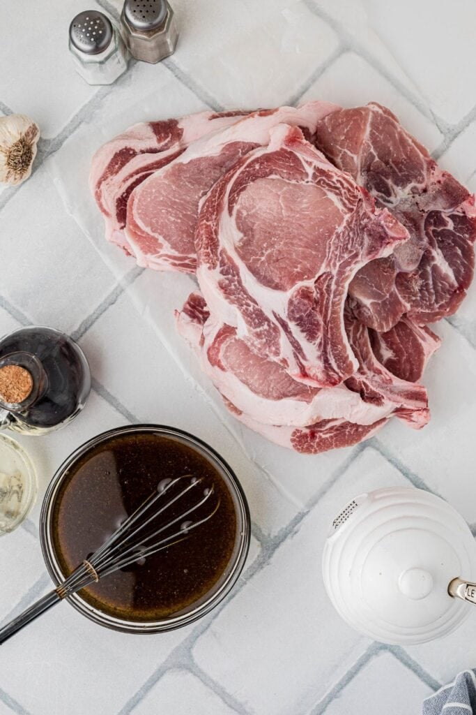 Raw bone-in pork chops in a pile next to a glass bowl of a homemade marinade.