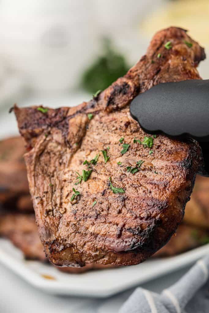 A pair of tongs lifting a grilled pork chop in front of a large serving platter of pork chops.