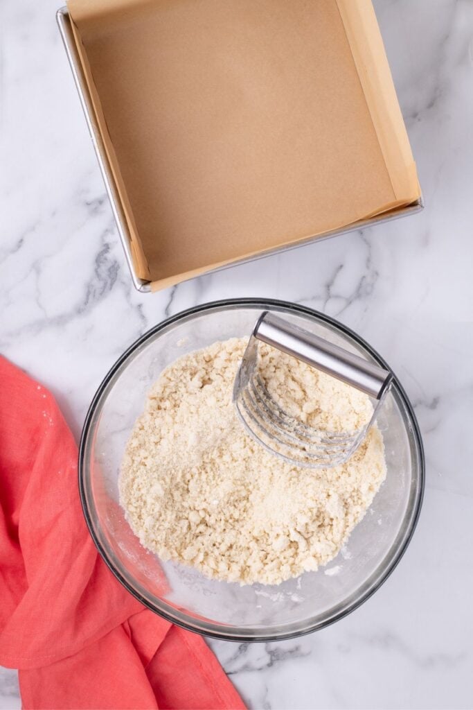 A glass bowl with ingredients for the crust and a pastry cutter in it, next to a parchment lined baking pan.