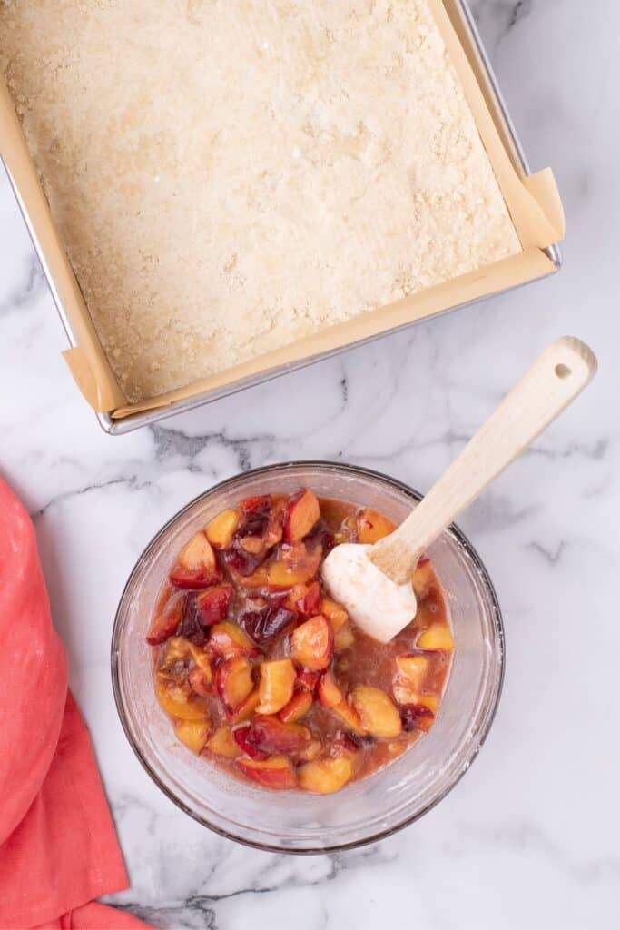 A glass bowl of peach filling next to a prepared crust in a parchment lined baking pan.