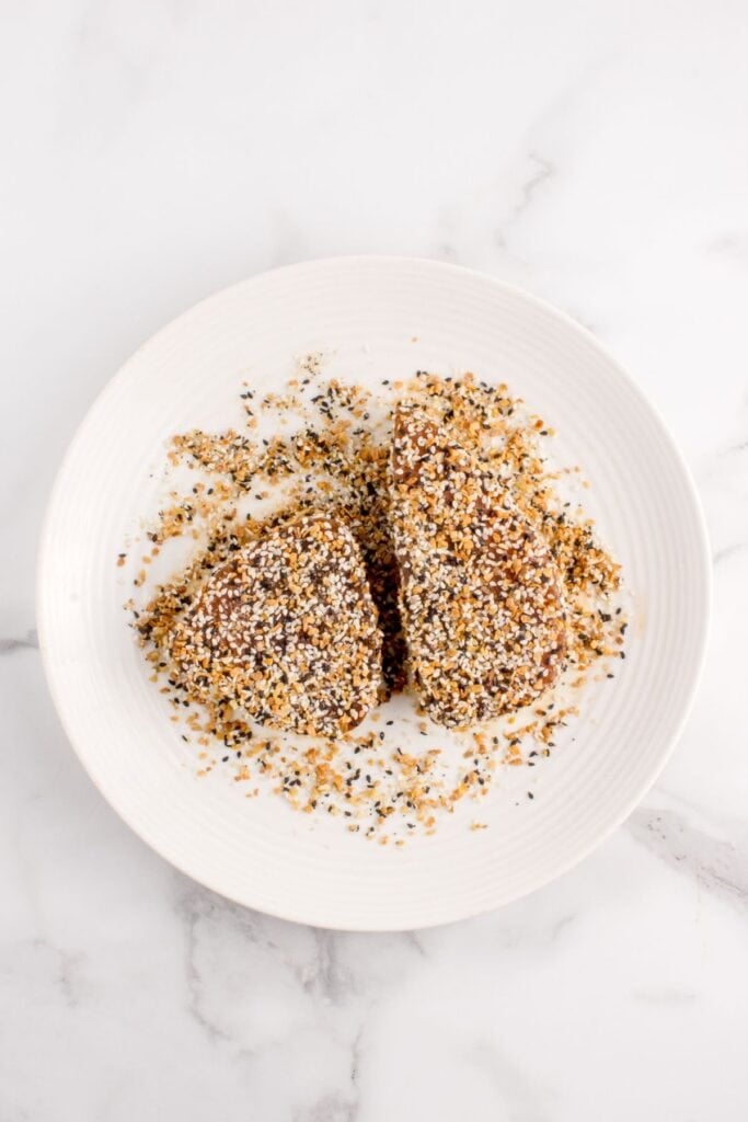 A white plate with two tuna steaks being dredged in everything bagel seasoning.