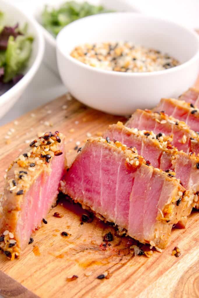 Sliced tuna steak on a wooden cutting board. A small bowl of seasoning is just behind the sliced tuna.