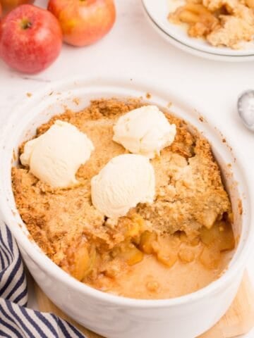 Baked apple crisp in a large white serving dish, with one serving removed and placed on a white plate behind it. Three scoops of ice cream sit on top of the crisp.