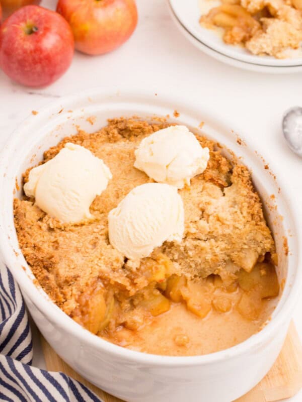 Baked apple crisp in a large white serving dish, with one serving removed and placed on a white plate behind it. Three scoops of ice cream sit on top of the crisp.