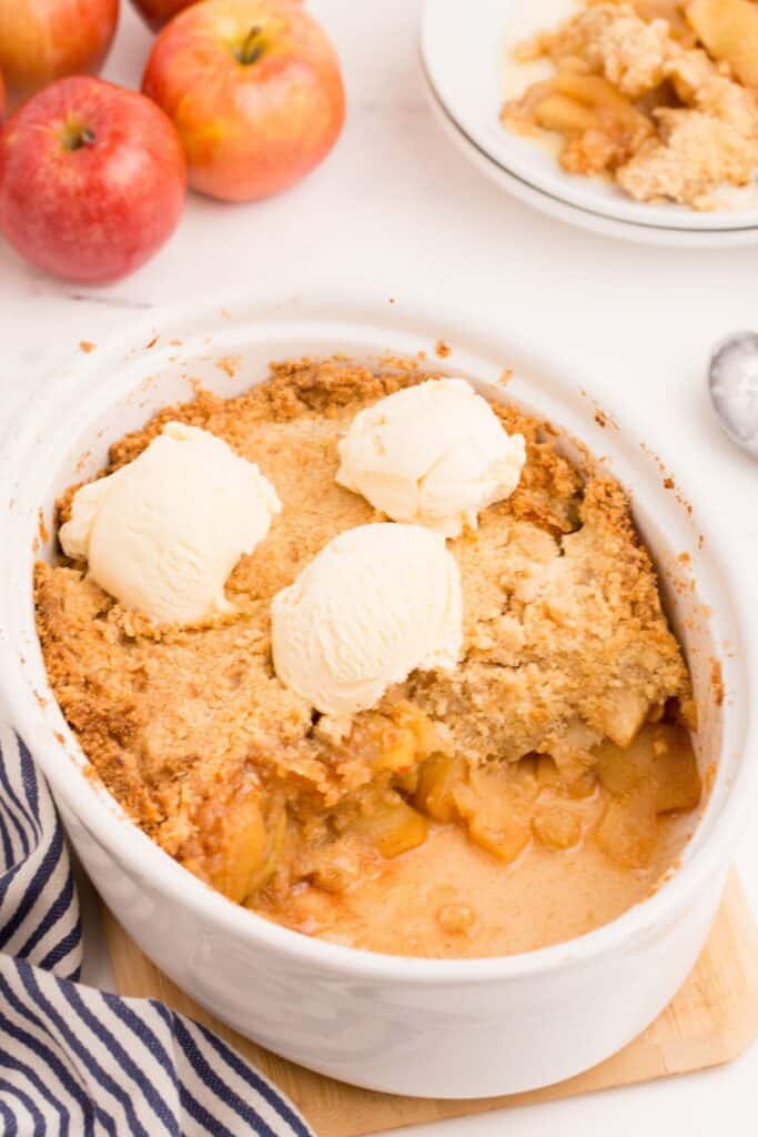 Baked apple crisp in a large white serving dish, with one serving removed and placed on a white plate behind it. Three scoops of ice cream sit on top of the crisp.