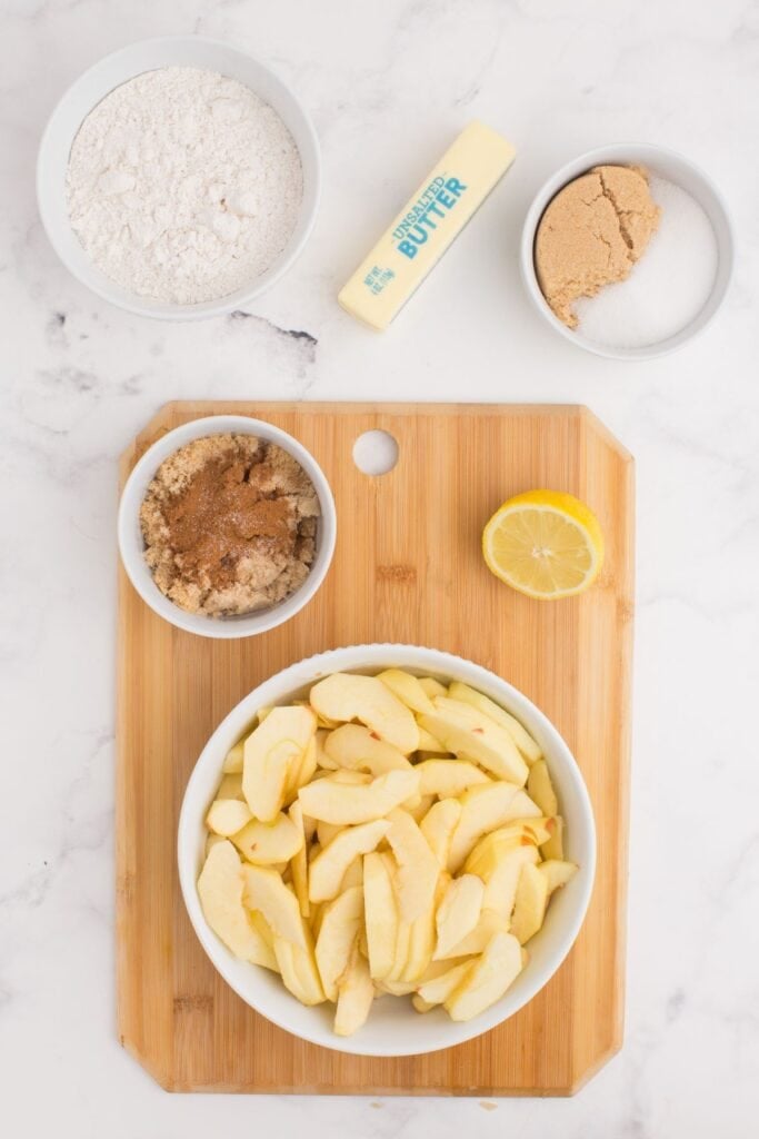 A wooden cutting board with a bowl of sliced apples, bowl of brown sugar and cinnamon, and half a lemon on it. Next to the cutting board is a bowl of flour, a stick of butter, and a bowl of brown sugar.