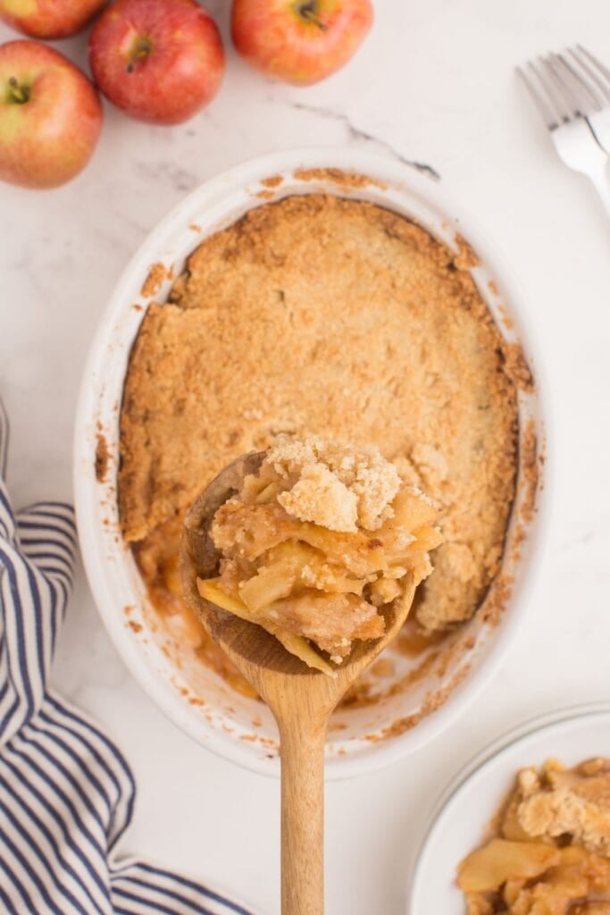 A wooden spoon is lifting a serving of apple crisp above the completed dessert.