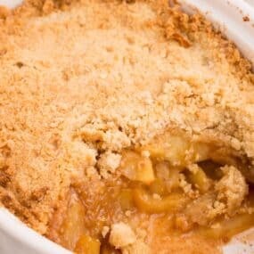 Completed apple crisp without oats in a white serving dish. A serving has been removed to show the baked apples inside.