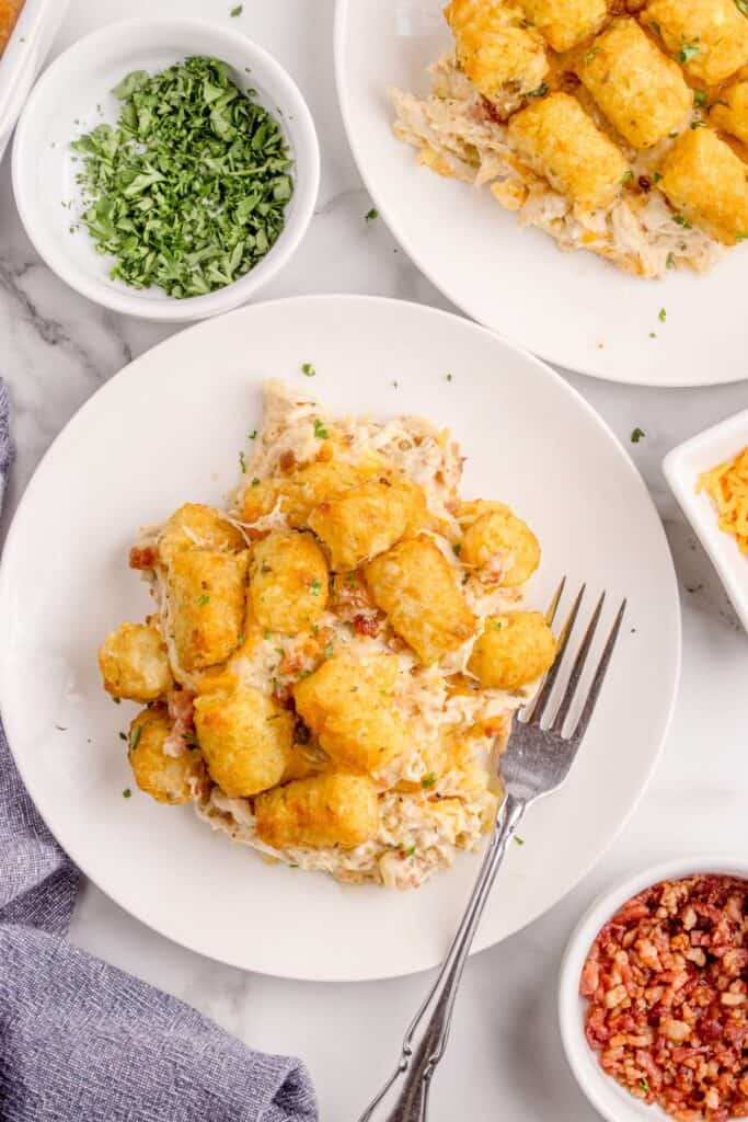 Casserole served on a white dinner plate surrounded by small bowls of bacon, cheese, parsley, and another dinner plate.