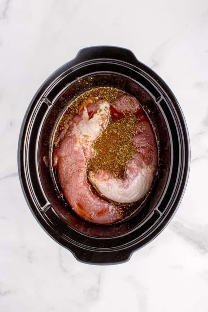 Raw pork tenderloin in a crock pot insert with juices and spices for cooking.