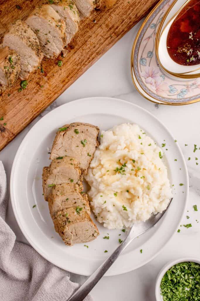 A white dinner plate with sliced pork tenderloin and mashed potatoes on it. Plate is surrounded by a small bowl of chopped parsley, a bowl of gravy, and a wooden cutting board with pork tenderloin slices lined up on it.