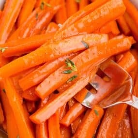 Maple glazed carrots in a white serving dish. A serving fork is holding a serving of carrots.
