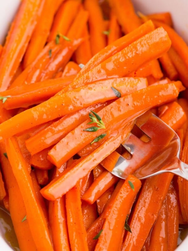 Maple glazed carrots in a white serving dish. A serving fork is holding a serving of carrots.