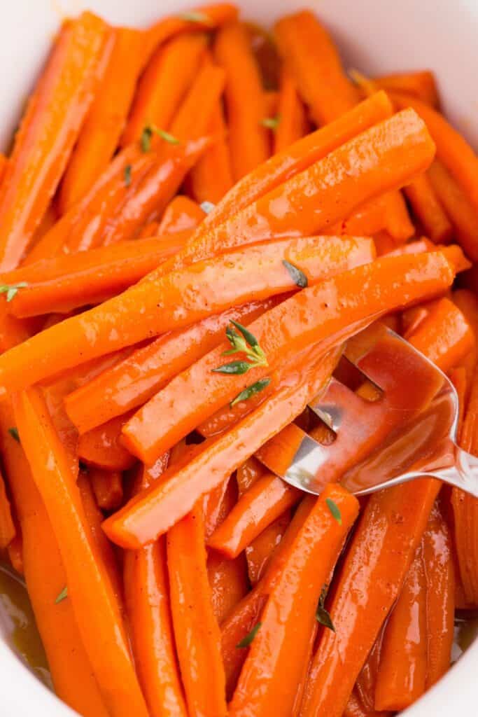Maple glazed carrots in a white serving dish. A serving fork is holding a serving of carrots.
