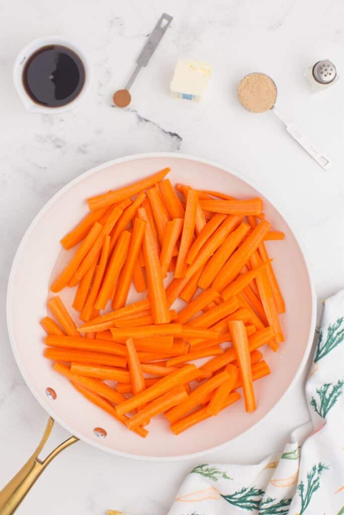 Peeled and chopped carrots placed in a skillet with water. Ingredients for the maple glaze are placed around the skillet.