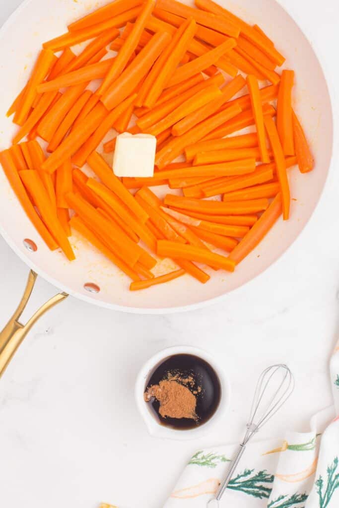Steamed carrots in a skillet with a large pat of butter on top. Next to the skillet is a small cup of maple syrup with cinnamon and brown sugar.