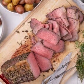 View of a partially sliced bottom round roast on a larger cutting board, flanked by serving bowls of baby potatoes and peas.