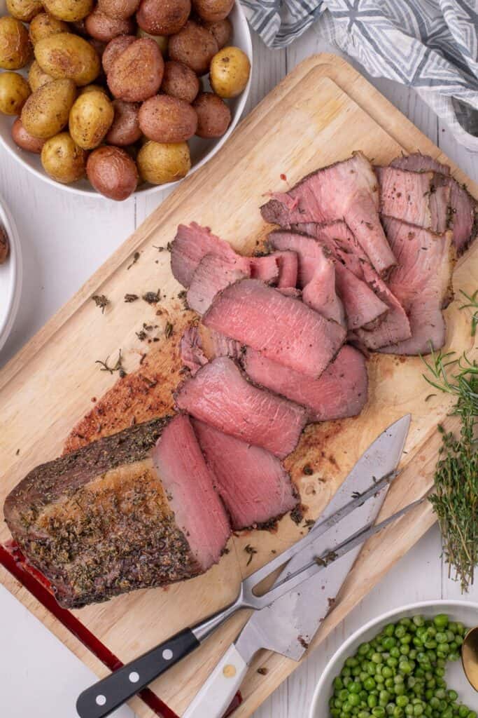 View of a partially sliced bottom round roast on a larger cutting board, flanked by serving bowls of baby potatoes and peas.